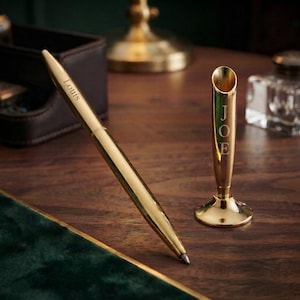 May include: A gold-toned metal pen engraved with the name Louis and a matching gold-toned metal pen stand engraved with the name JOE. The pen is positioned diagonally next to the upright cylindrical stand on a dark wooden surface.