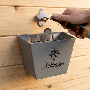 May include: A wall-mounted gray metal bottle opener and a matching gray metal bin for collecting bottle caps. The bin features a black star graphic and the text Kittridge printed on the front. A brown glass bottle is positioned at the opener.