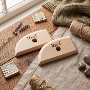 May include: Two semi-circular light-colored wooden weaving tools, each featuring a central circular hole and the name Leah engraved in a dark script font on the flat surface. The tools have a smooth, polished finish and a tapered edge.