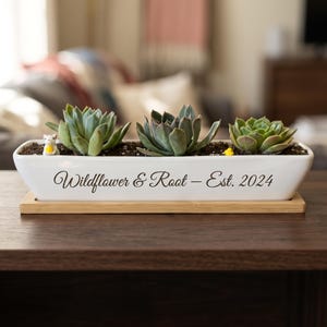 May include: A rectangular white ceramic planter with a bamboo base containing three green succulents. The front of the planter features the black text "Wildflower & Root – Est. 2024" and includes a small white mouse figurine and a tiny yellow bird.