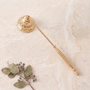 May include: A gold-toned metal candle snuffer with a bell-shaped head featuring the engraved name Céleste. The long, slender handle has a textured, knurled grip section and a rounded end, designed for extinguishing candle flames safely.