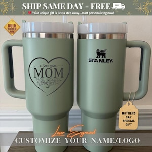 May include: Two light green Stanley tumblers with white lids and handles. One tumbler has "i love you MOM" in a heart with flowers. The other has the Stanley logo. Text: "Laser Engraved" and "Customize your name/logo". A tag says "Mothers Day Special Gift".