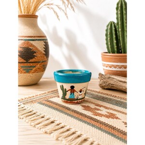 Op de afbeelding: Een kleine, crèmekleurige keramische plantenpot met een fel turquoise rand en een handgeschilderde scène van twee figuren die elkaars hand vasthouden tussen groene cactussen. De pot heeft een taps toelopende vorm en staat op een geweven mat.