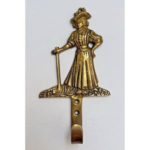 Puede incluir: Un gancho de pared de latón que representa a una mujer victoriana con un vestido largo y un sombrero, sosteniendo un bastón. El gancho está unido a una placa rectangular con dos orificios para tornillos y un gancho curvo en la parte inferior.