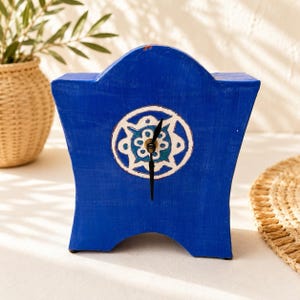 May include: A vibrant blue mantle clock with a flared, arched silhouette. The center features a circular white and teal floral motif surrounding black clock hands. The surface has a visible, hand-painted texture with a matte finish throughout.
