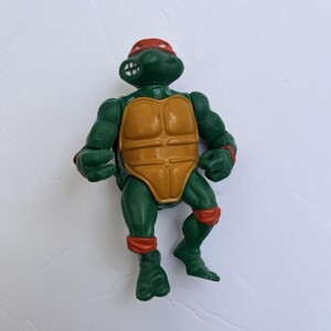 Puede incluir: Una figura de acción de plástico verde de un personaje de tortuga con complexión musculosa, que lleva un antifaz rojo, muñequeras rojas y bandas rojas en las piernas. La figura presenta un peto amarillo anaranjado y expresión decidida.