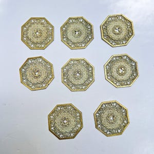Peut inclure: Huit dessous de verre octogonaux avec une bordure dorée et un motif de filigrane crème. Les dessous de verre présentent un motif central en forme d'étoile et sont disposés sur une surface blanche. Ils sont probablement décoratifs.