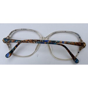 Puede incluir: Gafas con montura transparente y patillas estampadas en azul, marrón y dorado. Las gafas tienen una forma clásica y de gran tamaño, sobre un fondo blanco.