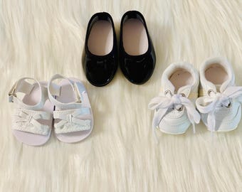 May include: Three pairs of shoes for a doll. The first pair is black patent leather flats. The second pair is white sandals with a woven design. The third pair is white sneakers with laces.