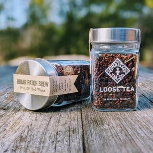 May include: Two glass jars of loose leaf tea with silver lids on a wooden surface. One jar is labeled "Briar Patch Brew Fruit & Herb Tisane." The other jar is labeled "Piper & Leaf Tea Co. Loose Tea" with 70 grams of tea.