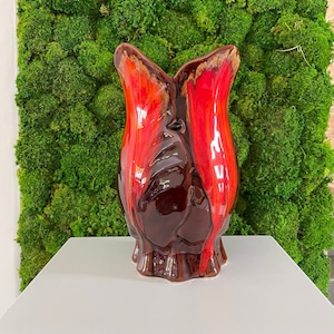 Peut inclure: Un vase en céramique rouge et marron avec un design abstrait unique. Le vase est posé sur une surface blanche devant un mur de mousse verte.