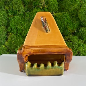 May include: A ceramic ashtray shaped like a house with a brown and green glaze. The ashtray has a small handle on the top and a wavy edge on the front.