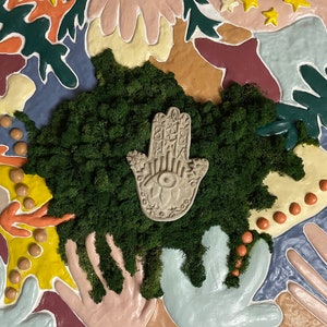 May include: Abstract wall art with a central patch of green moss and a beige Hamsa hand design. The background features shapes in orange, yellow, blue, and green. The artwork is a vibrant, colourful piece.