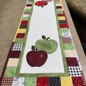 May include: A rectangular quilted table runner featuring a white center panel with appliqued red and green apple shapes. The border consists of a patchwork of small squares in various colors and patterns, including red, green, yellow, and black and white.