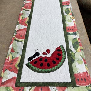 May include: A rectangular quilted table runner featuring a white center panel with two appliqued watermelon slices and black ants. The border is a dark green fabric, surrounded by a printed outer edge displaying red watermelon slices on white.