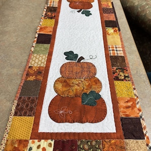 May include: A rectangular quilted table runner featuring a white center panel with two stacked pumpkin appliqués in shades of orange and brown. The border consists of a patchwork of various autumn-themed fabric squares in brown, gold, and orange tones.
