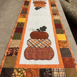 May include: A quilted table runner featuring a stacked pumpkin design. The pumpkins are in shades of orange and brown, with a plaid pattern on the middle pumpkin. The runner has a white center and is bordered with orange and various fall-themed fabric squares.