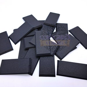 Op de afbeelding: Een stapel van 15 zwarte rechthoekige plastic sokkels voor miniatuurfiguren. De sokkels zijn getextureerd en hebben een verhoogde rand. De sokkels zijn in een willekeurig patroon gerangschikt.