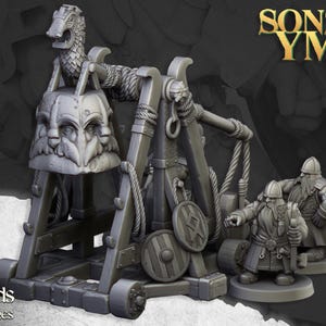 Puede incluir: Un modelo en miniatura de plástico gris de una catapulta con tres enanos. La catapulta tiene una cuerda y una cabeza grande de tres caras en la parte delantera. Los enanos sostienen armas y escudos. El texto "Sons of Ymir" está en el fondo.
