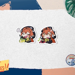 Puede incluir: Dos pegatinas con un personaje de dibujos animados con pelo naranja y un atuendo negro. Una pegatina muestra al personaje con un bocadillo y comida, la otra con dinero. Las pegatinas están sobre un fondo blanco con una pancarta amarilla que dice "WITH FREEBIES!!".