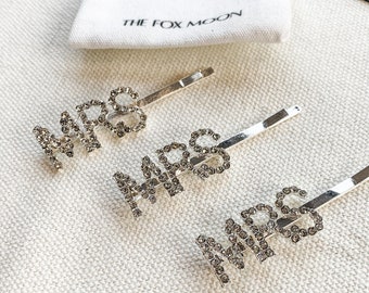 May include: Three silver hair clips with the word "MRS" spelled out in rhinestones. The clips are on a white fabric background with the text "THE FOX MOON" printed on a white pouch.