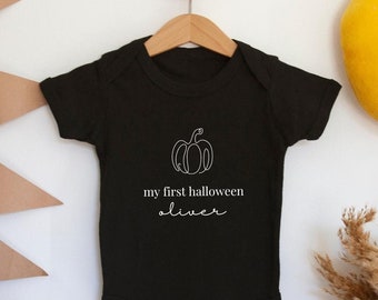 May include: Black baby onesie with a white pumpkin and the text "my first halloween oliver" on the front. The onesie is hanging on a wooden hanger against a white wall.
