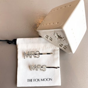 K&ouml;nnte beinhalten: Zwei silberne Haarklammern mit dem Wort "MRS" in Strasssteinen. Die Klammern befinden sich in einem wei&szlig;en Kordelbeutel mit dem Aufdruck "THE FOX MOON" auf der Vorderseite.