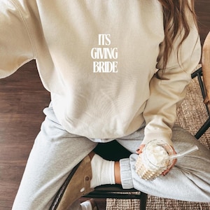 Könnte beinhalten: Beiges Sweatshirt mit dem Text "ITS GIVING BRIDE" in Weiß. Die Person trägt eine graue Jogginghose, weiße Socken und braune Sandalen. Sie hält ein Glas Eiskaffee.