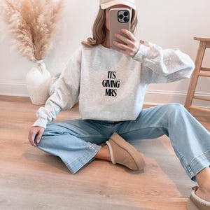 Könnte beinhalten: Ein hellgraues Sweatshirt mit dem Text "ITS GIVING MRS" in Schwarz. Die Person trägt eine hellblaue Jeans, beige Schuhe und eine weiße Baseballkappe. Im Hintergrund befindet sich eine Vase mit getrocknetem Pampasgras.