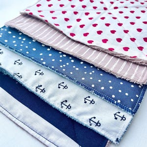 Può includere: Una pila di quadrati di tessuto trapuntato con vari motivi. Il quadrato superiore ha uno sfondo bianco con cuori rossi. Sotto c'è un quadrato a righe rosa e bianco, seguito da un quadrato blu con pois bianchi. I quadrati inferiori presentano motivi di ancore e strisce.