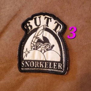 Puede incluir: Parche bordado en blanco y negro con el texto "BUTT SNORKELER" y un gr&aacute;fico de una persona con un snorkel y equipo de buceo. El parche tiene un borde negro y est&aacute; cosido sobre una tela marr&oacute;n.
