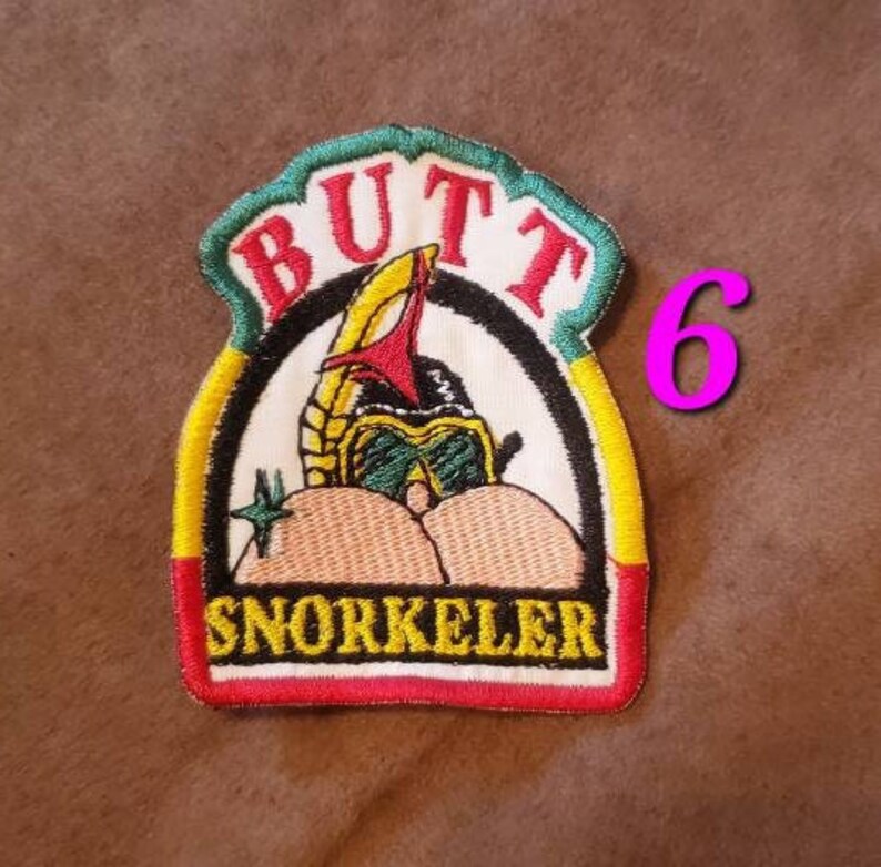 Puede incluir: Un parche bordado en verde, amarillo y rojo con las palabras "Butt Snorkeler" y una imagen de dibujos animados de una persona con un snorkel y una m&aacute;scara de buceo. El n&uacute;mero "6" est&aacute; escrito en rosa en la esquina superior derecha de la imagen.