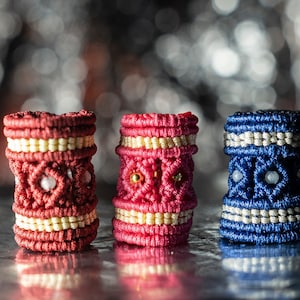 May include: Three cylindrical macrame beads featuring intricate knotted patterns. The left bead is dark red with cream accents, the centre bead is bright pink with cream and gold details, and the right bead is deep blue with silver accents.