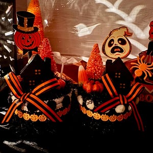 May include: Two round black containers decorated with orange and black striped ribbons, featuring miniature black houses, orange bottle brush trees, small pumpkins, and candy corn. Toppers include a pumpkin in a top hat and a ghost figure.