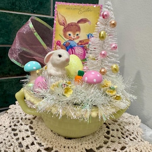 May include: A vintage Easter centerpiece in a pale yellow teapot. It features a bunny figurine, a greeting card, a small white Christmas tree, and decorative eggs and mushrooms. The arrangement sits on a crocheted doily.