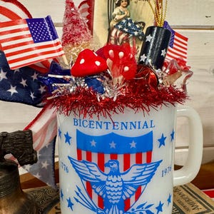 May include: A white mug with a patriotic theme, featuring a blue eagle emblem and the word "BICENTENNIAL". The mug is decorated with miniature American flags, red and white tinsel, and a vintage-style image of a girl with a flag.