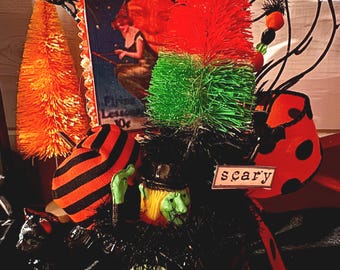 May include: A decorative Halloween display featuring a central witch figure with a green face, a black hat, and a striped orange and black cap. It includes a red and green bottle brush tree, a small sign reading scary, and a vintage-style witch illustration.