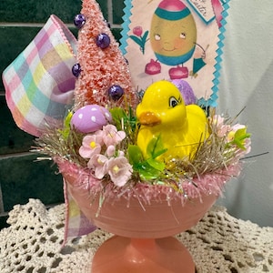 May include: A vintage Easter centerpiece in a pink pedestal bowl. It features a yellow duck, a pink bottle brush tree, purple eggs, and a greeting card that says "Happy Easter". The arrangement is decorated with pink flowers and a pastel ribbon.