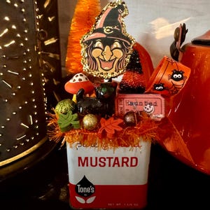 May include: A vintage Tone's mustard tin filled with Halloween decorations including a witch cutout with gold glitter edges, a black cat figurine, a mushroom, orange bottle brush trees, a spider ribbon, and a ticket labeled Haunted.