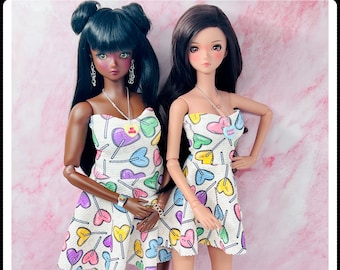 May include: Two dolls wearing white dresses with a lollipop heart print. The dresses have a strapless design and are accented with colorful hearts and lollipops. The dolls are wearing silver heels and have long black hair. One doll has brown skin and the other has fair skin.