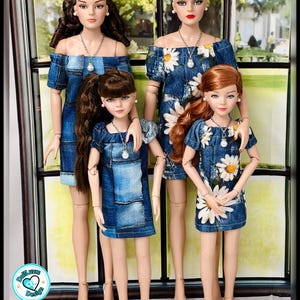 May include: Four fashion dolls in denim dresses. Two dolls wear off-the-shoulder dresses, one with a daisy print. The other two dolls wear denim dresses with a patchwork design. Each doll wears a silver necklace with a pearl pendant. The dolls are standing in front of a window.