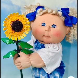 May include: A Cabbage Patch Kid doll with blonde curly hair wearing a blue and white tie-dye dress and white sweater, holding a yellow Lego sunflower.