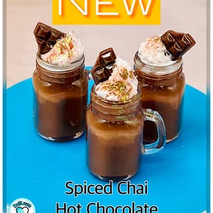 May include: Three small glass jars filled with a brown liquid topped with whipped cream and a chocolate bar. The jars are on a blue surface and the text "Spiced Chai Hot Chocolate Small" is below the jars.