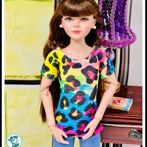 May include: A doll wearing a colorful leopard print top with blue jeans. The top features a vibrant mix of yellow, blue, pink, and black spots. The doll has long brown hair and is wearing pearl earrings.