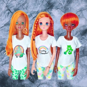 May include: Three dolls with orange hair wearing white t-shirts with different graphic designs. The dolls are wearing green and white patterned pants. The t-shirts say "Lucky Charm!" and "Shamrockin' the Day".