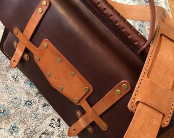 May include: A brown leather satchel with a tan leather strap and a brass buckle. The bag has a top handle and a flap closure.
