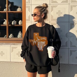 Puede incluir: Conjunto de sudadera y pantalones cortos negros. La sudadera presenta un gráfico marrón en forma de Texas con las palabras "Hook 'em Horns". El conjunto se usa con gafas de sol y un bolso negro.