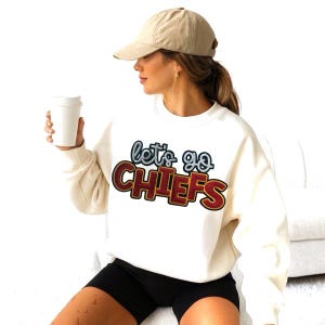 May include: Cream-colored sweatshirt with the text "Let's Go Chiefs" in a layered design. The text is outlined in blue and red, with a brown border. The person is wearing a tan baseball cap and black shorts.