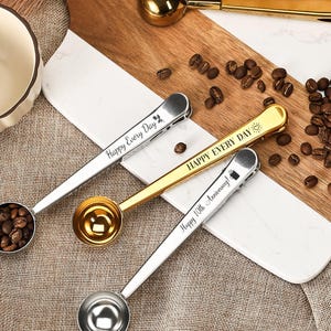 Puede incluir: Cucharas de café con clips para bolsas integrados en oro y plata. Las cucharas tienen grabado el texto "Happy Every Day" y "Happy 10th Anniversary". Granos de café esparcidos sobre una tabla de cortar de madera.