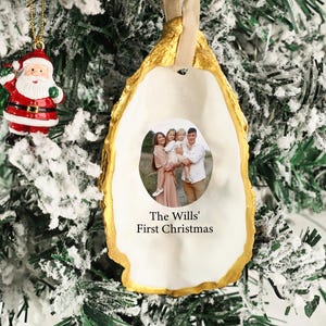 May include: A Christmas ornament shaped like an oyster shell, with a gold-painted edge and a family photo in the center. The text "The Wills' First Christmas" is printed below the photo. A small Santa Claus ornament hangs nearby.